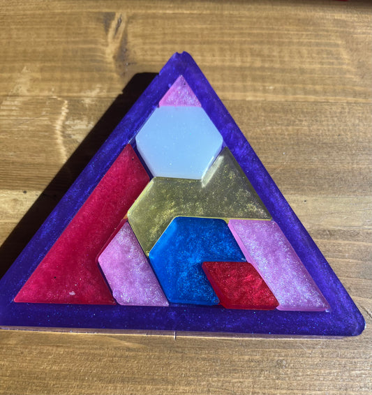 A triangular puzzle made of resin with a purple border and colourful geometric pieces inside. The puzzle pieces are in shades of pink, blue, gold, and white.