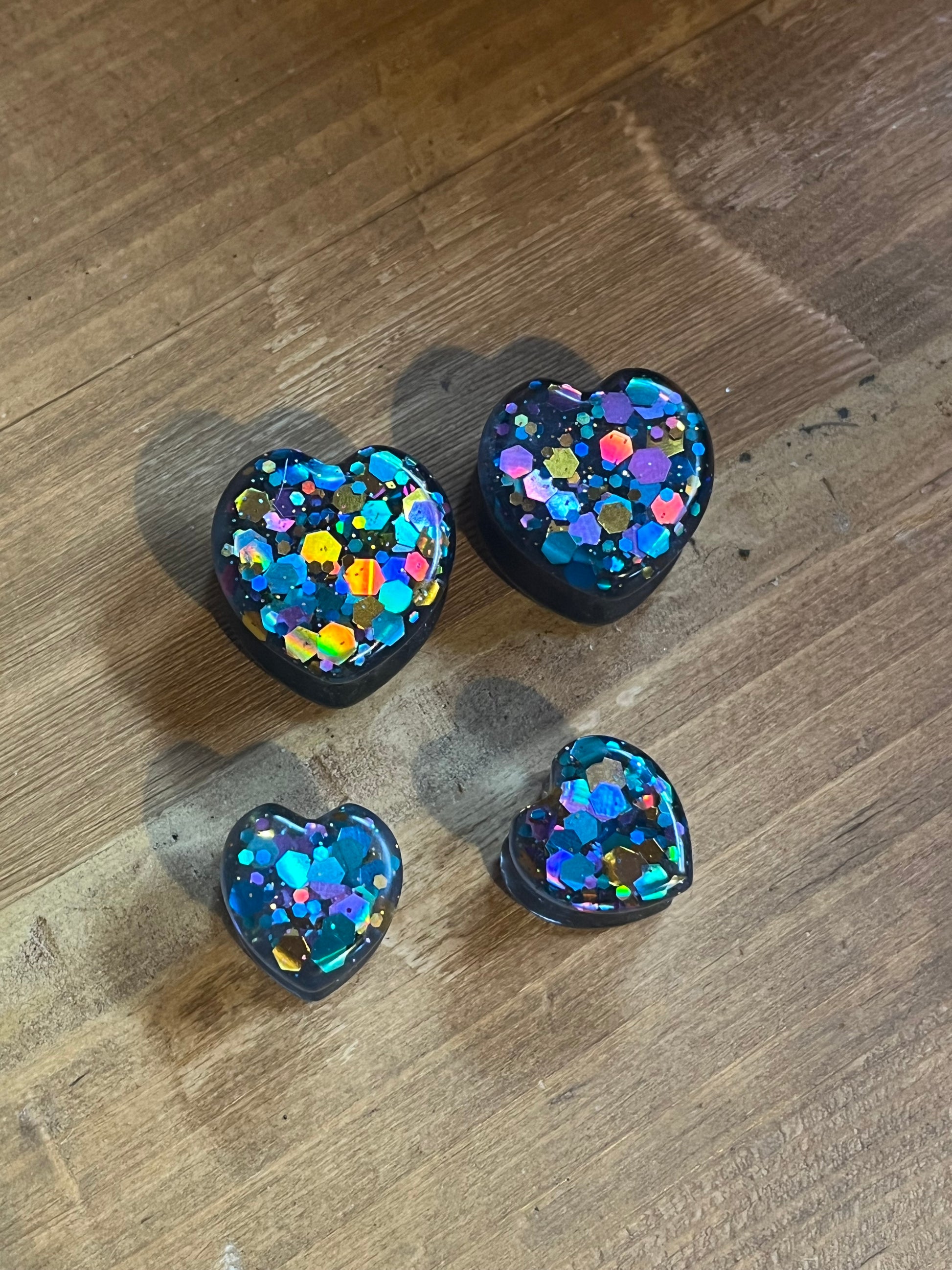 Four heart-shaped plugs with a black base and filled with colourful glitter. The glitter includes shades of blue, purple, gold, and pink. The plugs are arranged on a wooden surface.