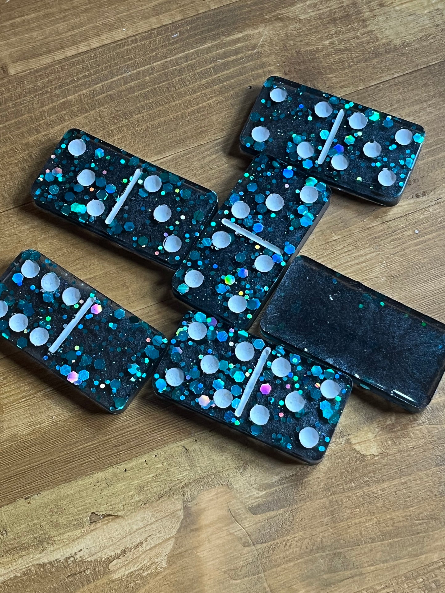 Six dominoes with a dark teal background and iridescent glitter. Each domino has white dots and a white dividing line. The dominoes are arranged on a wooden surface.