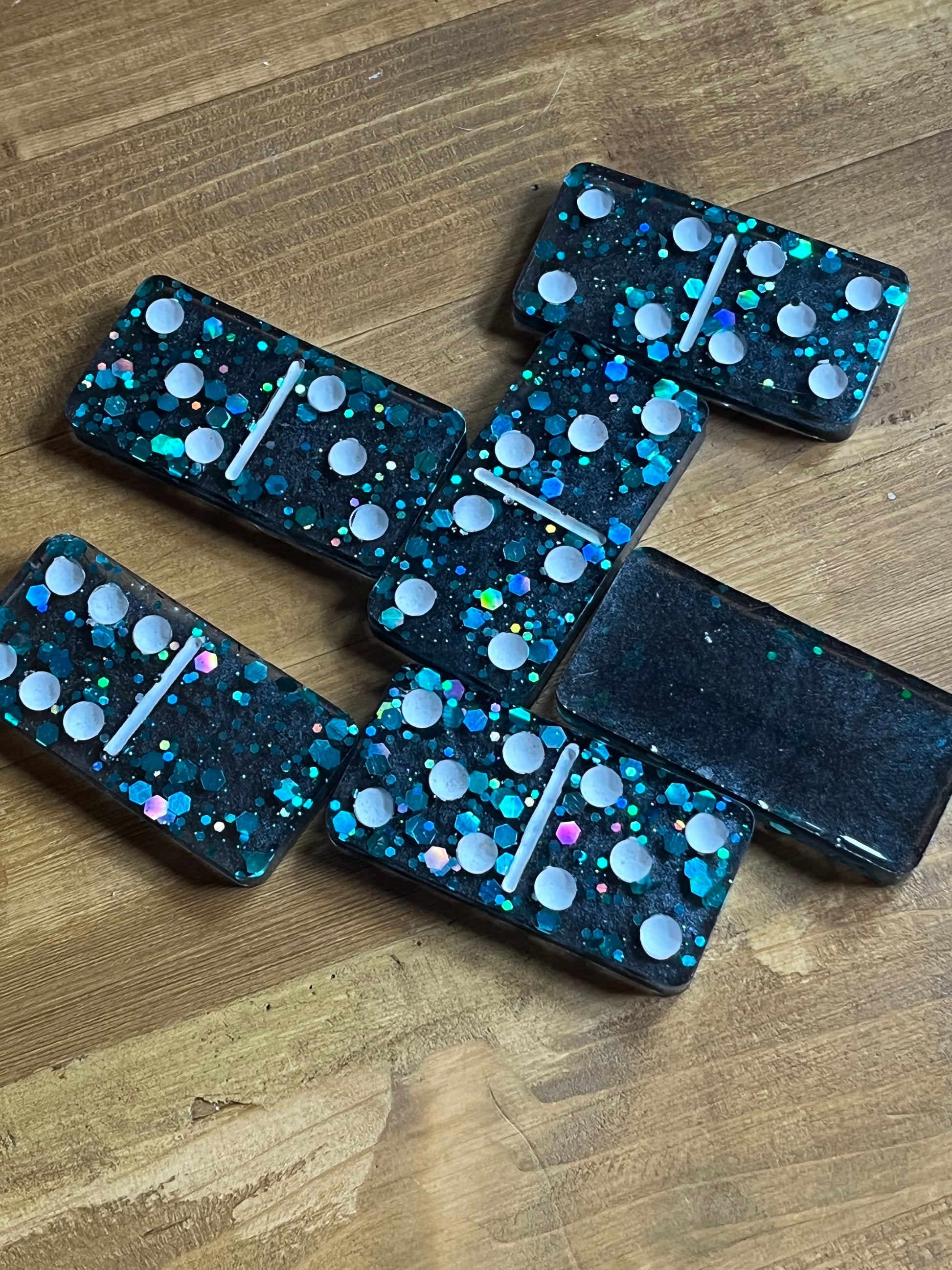 Six dominoes with a dark teal background and iridescent glitter. Each domino has white dots and a white dividing line. The dominoes are arranged on a wooden surface.