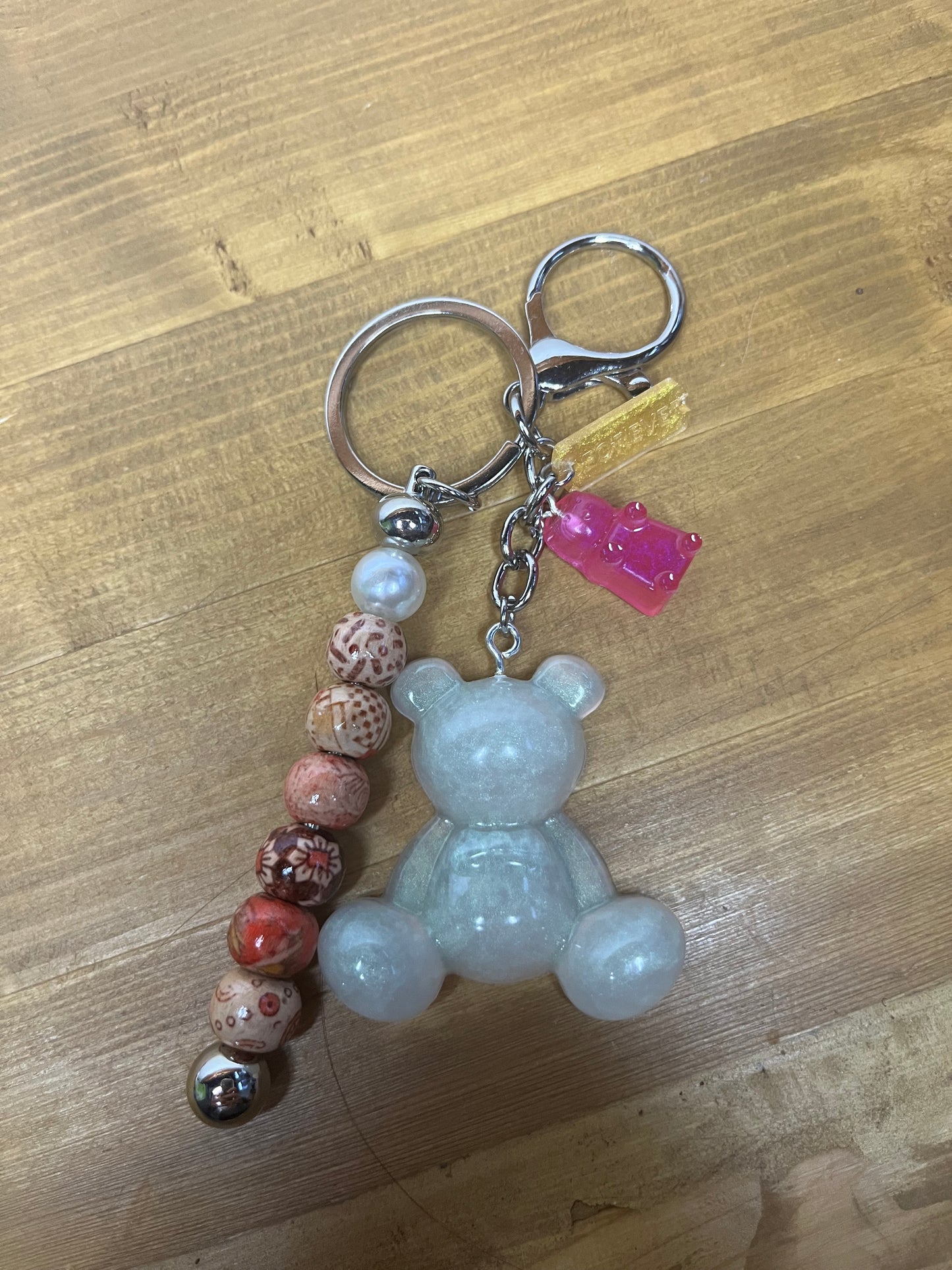 A silver keyring featuring a light grey teddy bear charm, a pink gummy bear charm, and a string of decorative beads in red, brown, and white. The keyring has a silver clasp and a key ring.