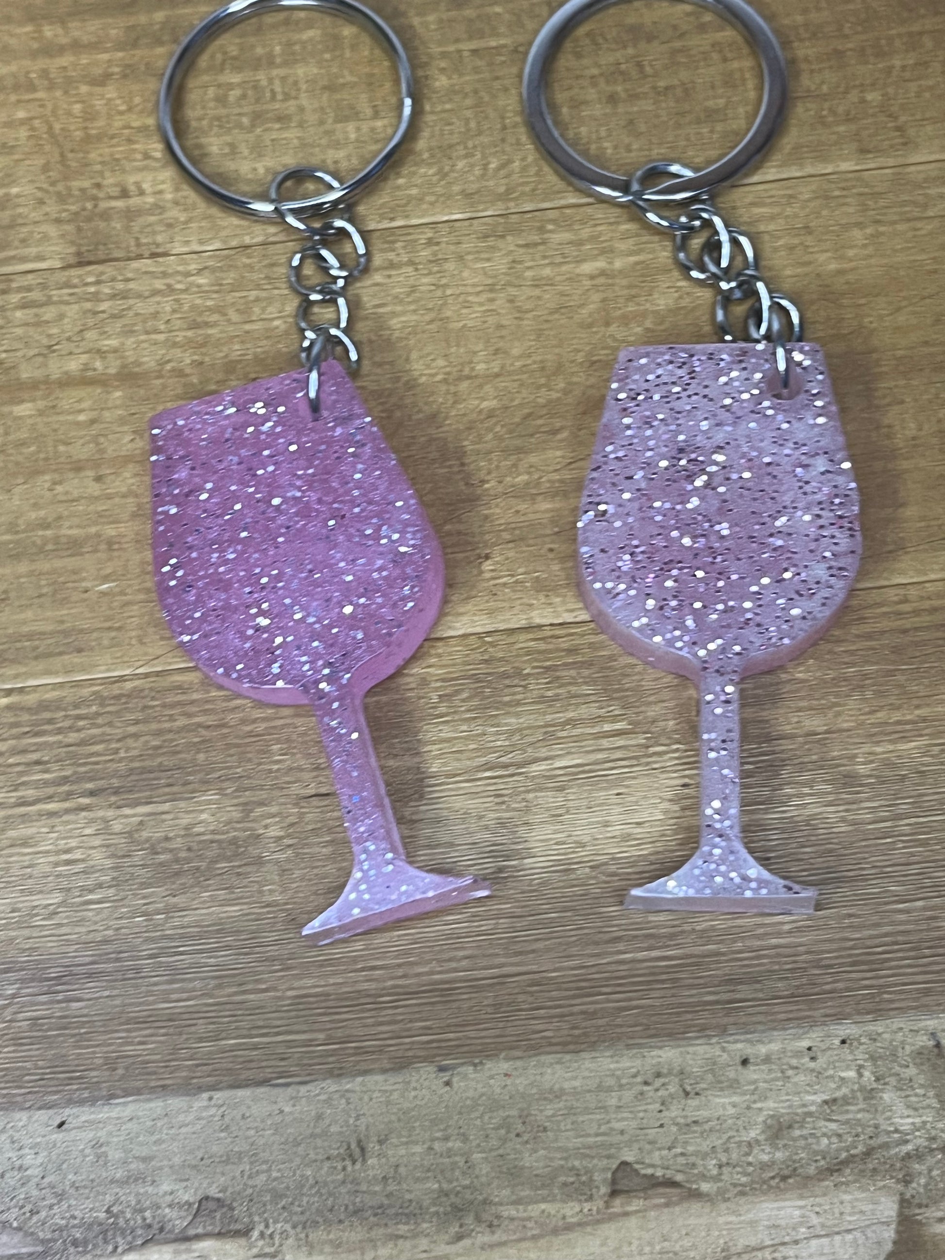 Two keyrings with a wine glass design. The wine glasses are a light pink colour and filled with silver glitter. Each keyring has a silver metal ring and chain.