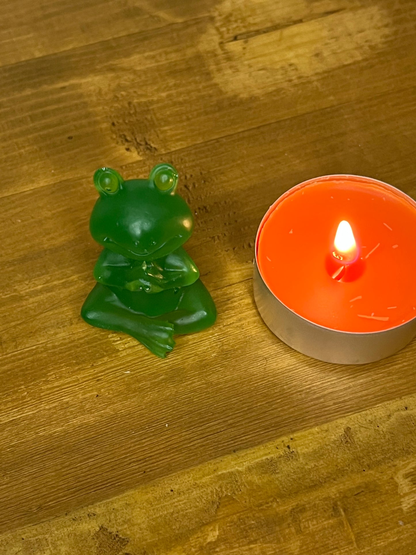 A green frog ornament in a seated lotus position beside a lit orange candle in a silver container. The frog is a solid, glossy green. The candle flame is bright and the background is a wooden surface.