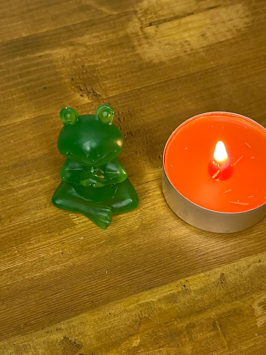 A green frog ornament in a seated lotus position beside a lit orange candle in a silver container. The frog is a solid, glossy green. The candle flame is bright and the background is a wooden surface.
