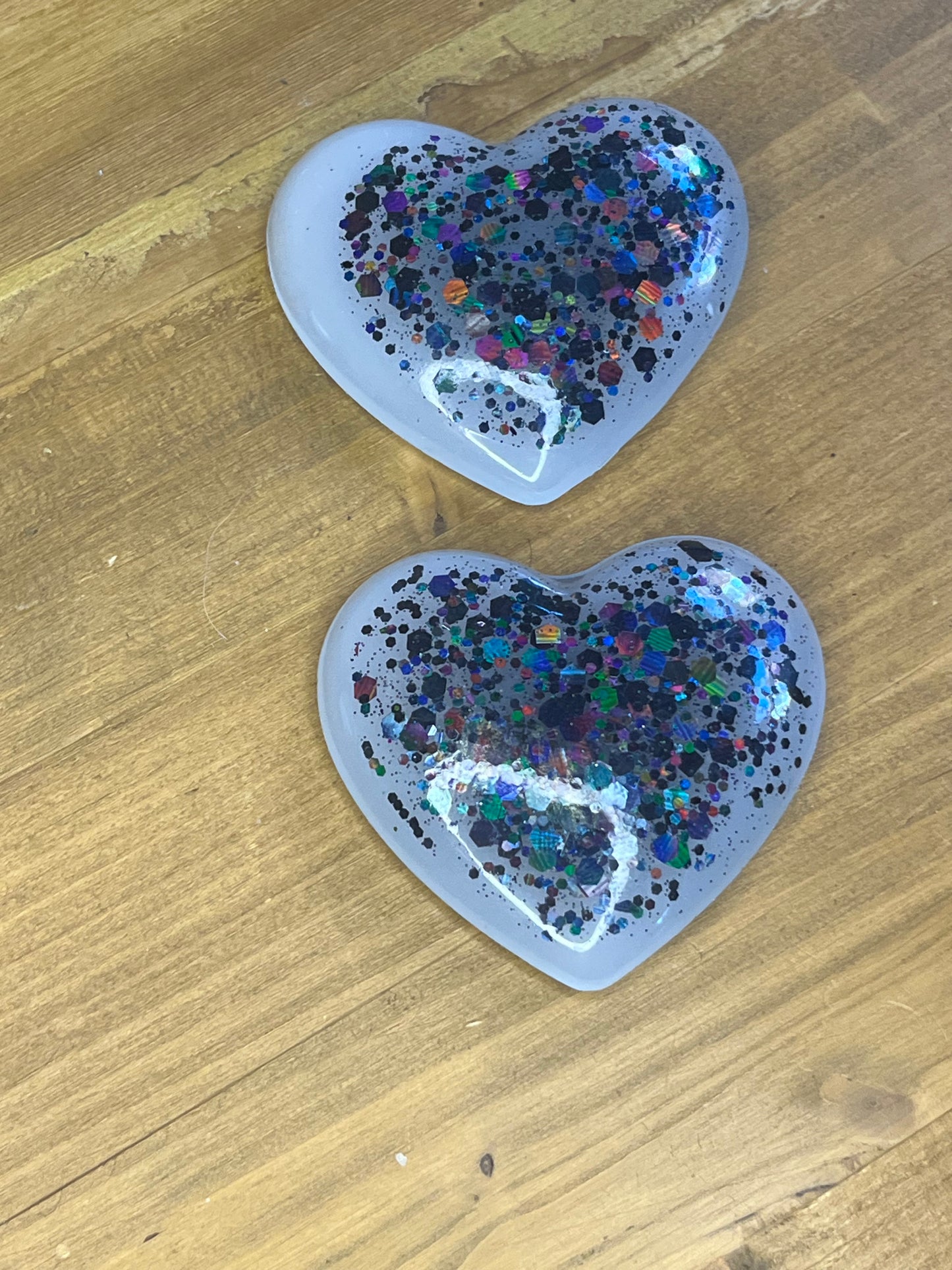 Two heart-shaped resin magnets. The magnets are white with a glittery, colourful design. The glitter includes shades of blue, purple, green, and silver. The magnets are displayed on a wooden surface.