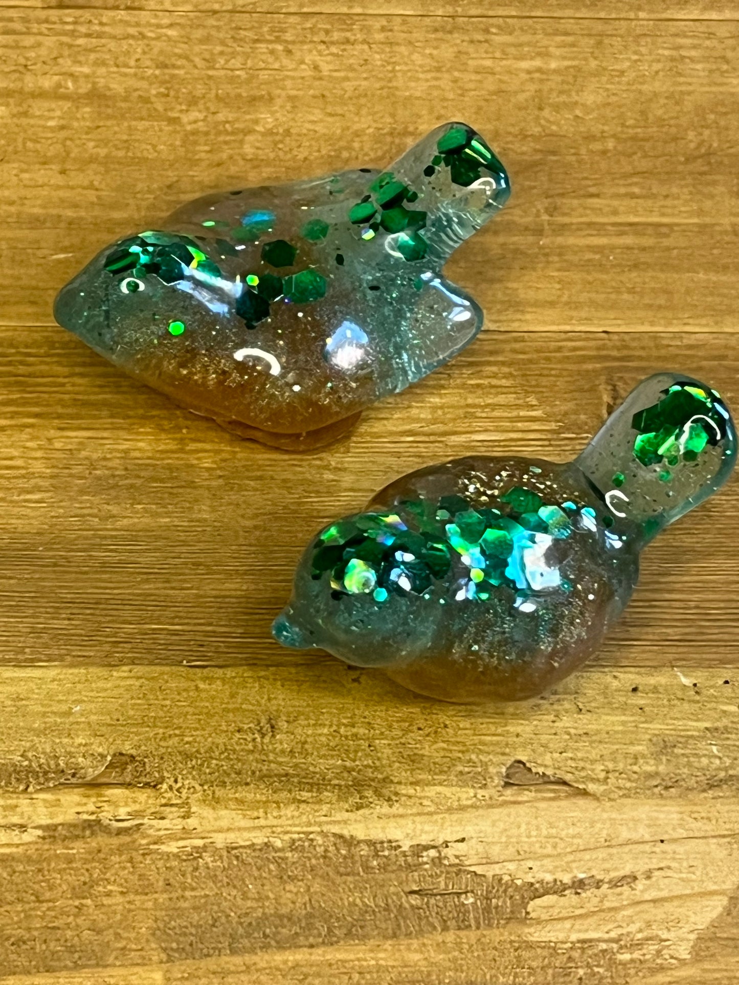 Two resin bird-shaped figurines. The birds are a mix of brown, blue, and green with green glitter embedded throughout. The birds are on a wooden surface.