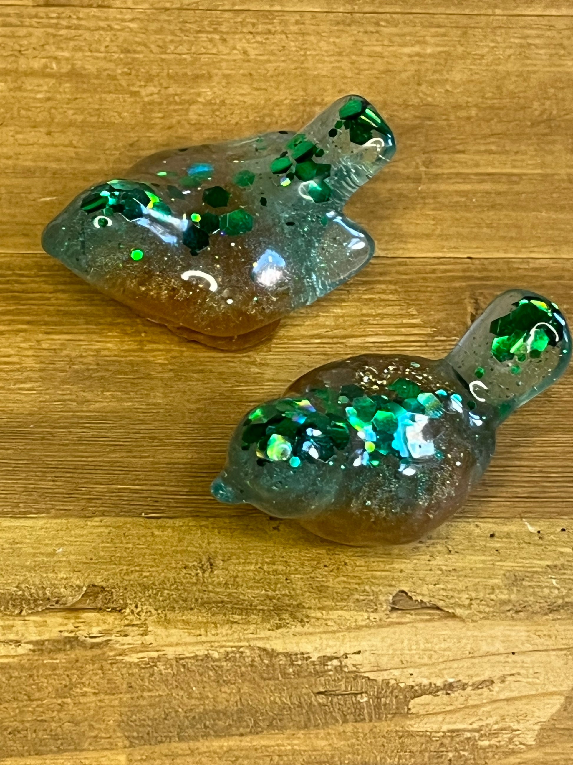 Two resin bird-shaped figurines. The birds are a mix of brown, blue, and green with green glitter embedded throughout. The birds are on a wooden surface.