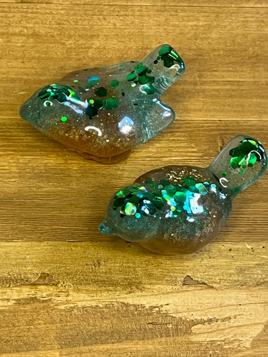 Two resin bird-shaped figurines. The birds are a mix of brown, blue, and green with green glitter embedded throughout. The birds are on a wooden surface.