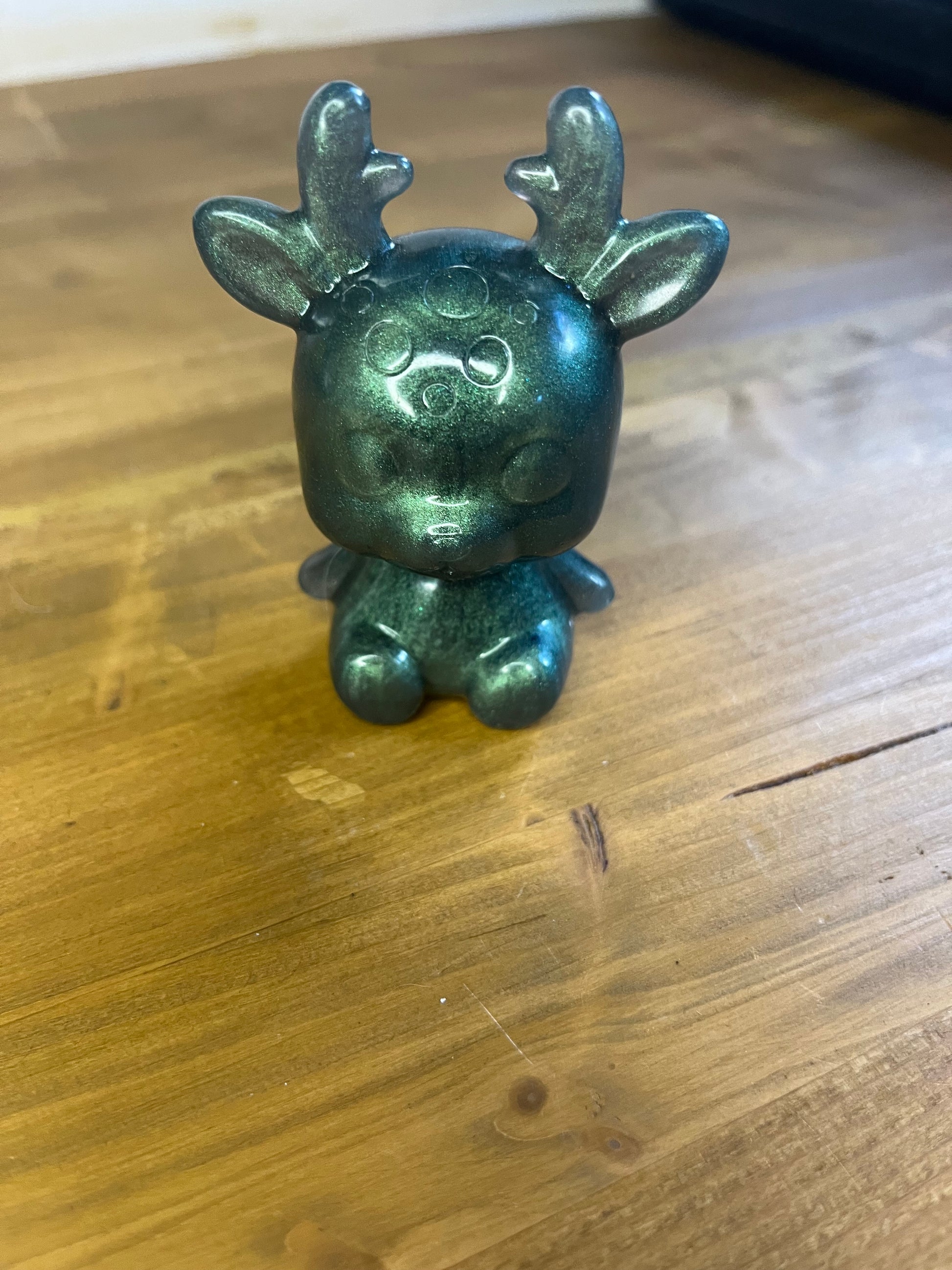 A small, stylized deer figurine with a metallic green finish. The deer has large, rounded eyes, small antlers, and a squat body. The figurine is sitting on a wooden surface.