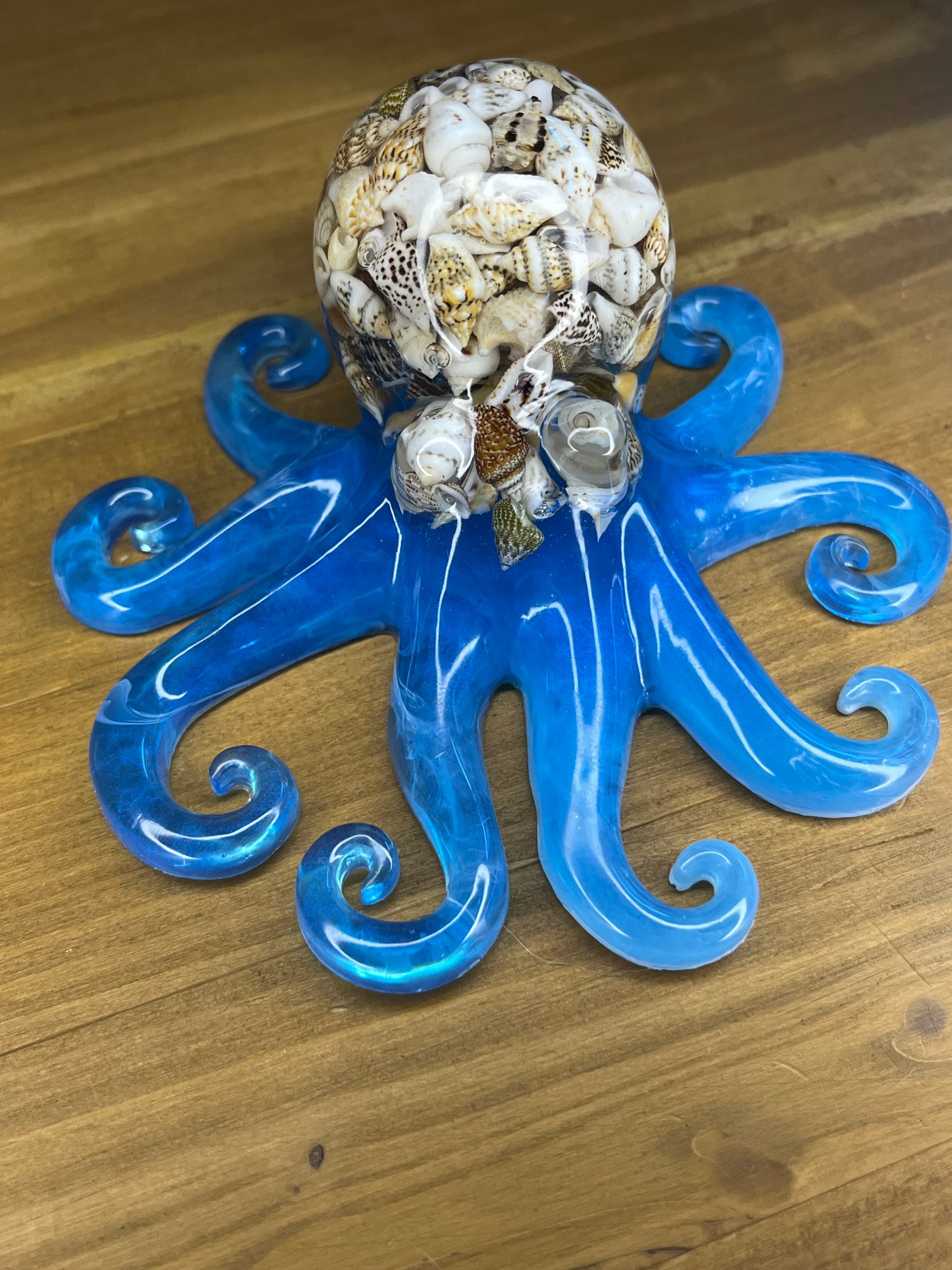 A decorative octopus sculpture. The body is made of blue resin, and the head is made of seashells. The octopus has eight curled tentacles. It is displayed on a wooden surface. A unique home decor item.