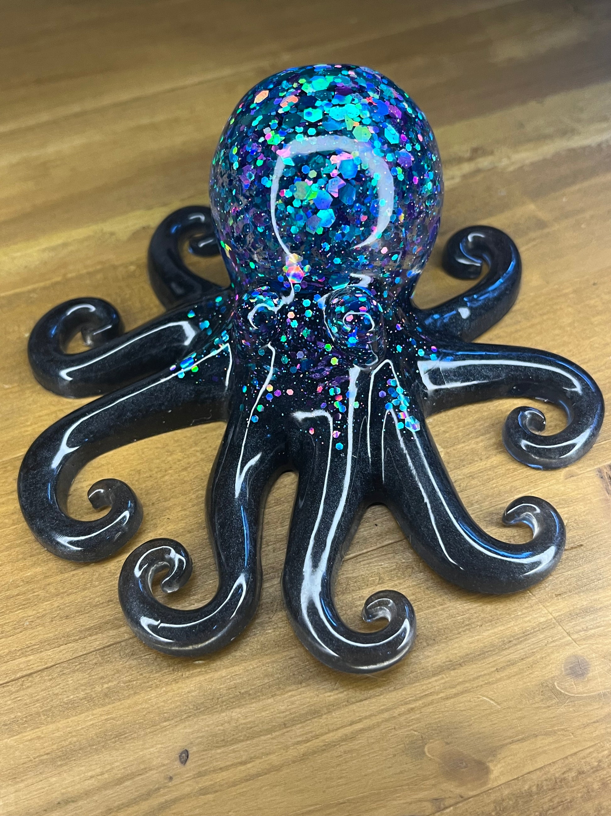 A decorative octopus figurine. The black body has eight curled tentacles. The head is a sphere filled with iridescent glitter in shades of blue, green, and purple. The figurine is set on a wooden surface.