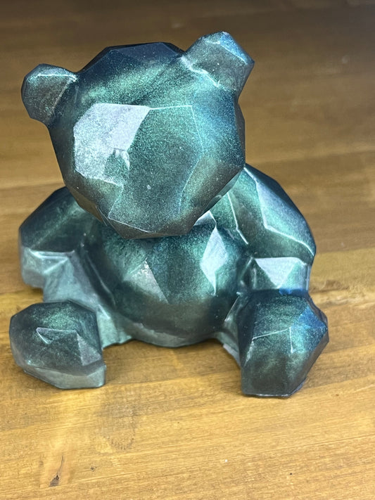A geometric bear figurine in green and blue hues. The bear is in a sitting position, with its body formed from numerous flat, angular facets. The figurine has a metallic finish and is placed on a wooden surface.