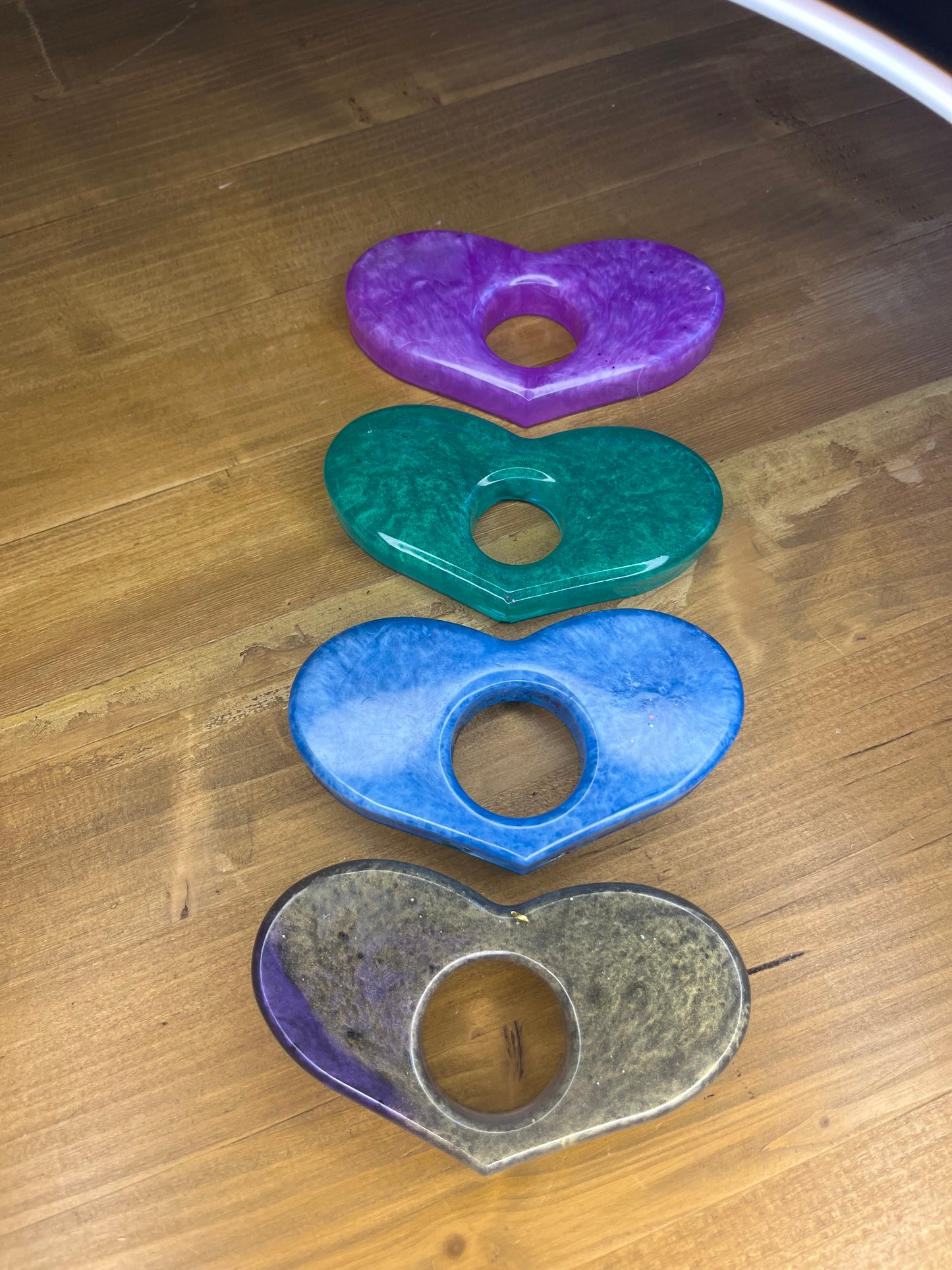 Four heart-shaped resin pieces in purple, teal, blue, and grey with a central hole. The pieces are arranged vertically on a wooden surface, showcasing a glossy finish and varying colour gradients. The design is simple and elegant.