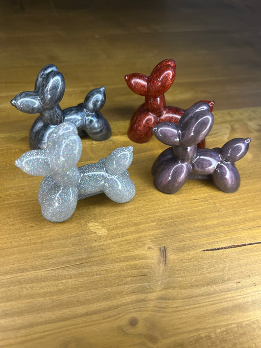 Four glittery balloon dog figurines in silver, red, grey, and purple. The dog sculptures are posed in a playful position, with their tails and ears up. The figurines are displayed on a wooden surface.