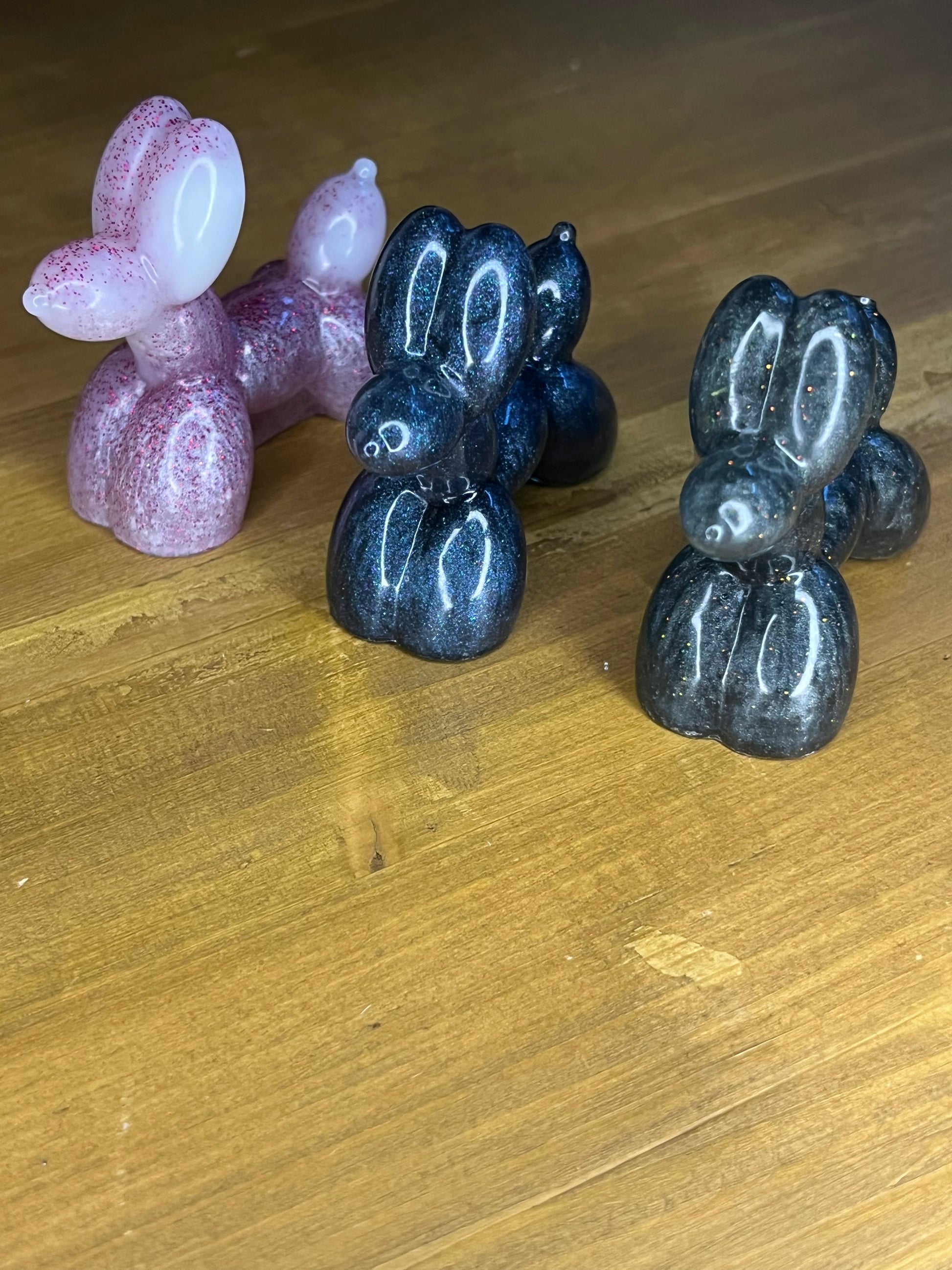Three resin balloon dog figurines in pink, navy blue, and black. The pink dog has glitter accents, while the navy and black dogs have a sparkly, iridescent finish. Each dog has a rounded body and head, with a small, protruding snout.