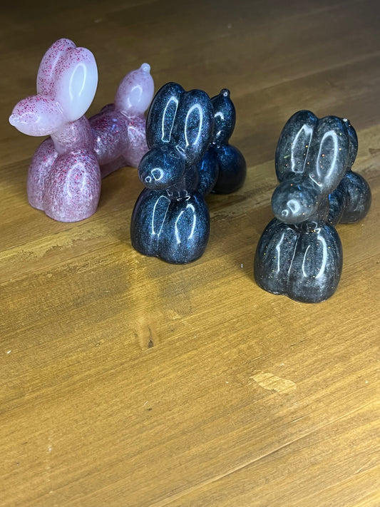 Three resin balloon dog figurines in pink, navy blue, and black. The pink dog has glitter accents, while the navy and black dogs have a sparkly, iridescent finish. Each dog has a rounded body and head, with a small, protruding snout.