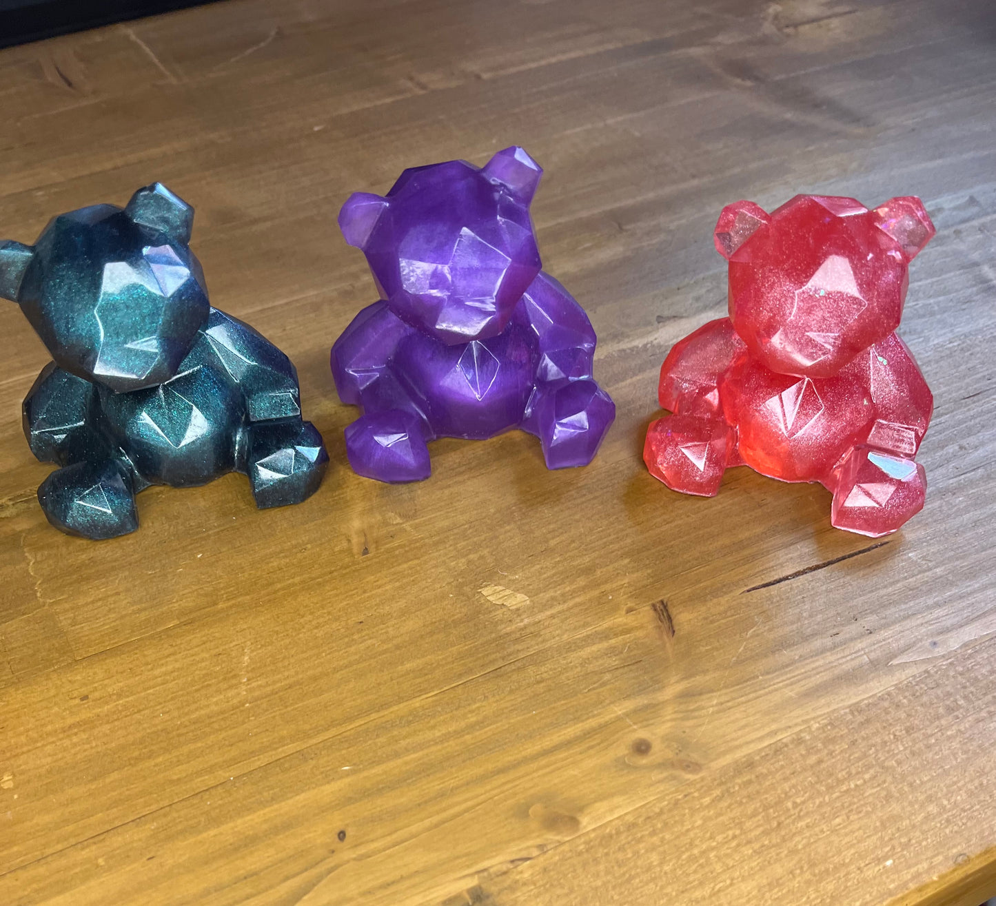Three faceted resin teddy bear figurines in dark green, red, and purple. The bears are sitting on a wooden surface. The bears have a geometric design.