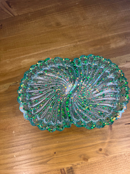 Transparent resin ornament shaped like two joined scallop shells, filled with green, gold, and multicoloured glitter, displayed on a wooden surface.