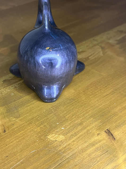 A small, decorative dolphin figurine in shades of purple and blue. The resin dolphin has a smooth, glossy finish and is posed with its tail and dorsal fin raised. The figurine is resting on a wooden surface.