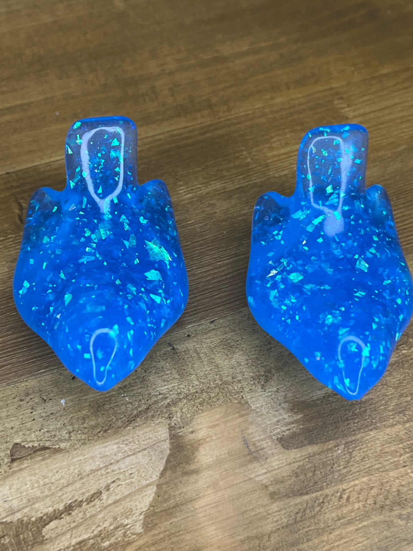 Two blue resin figurines with a teardrop shape and a textured, glittery appearance. They have a vibrant blue colour with embedded iridescent flakes, creating a sparkling effect. The background is a wooden surface.
