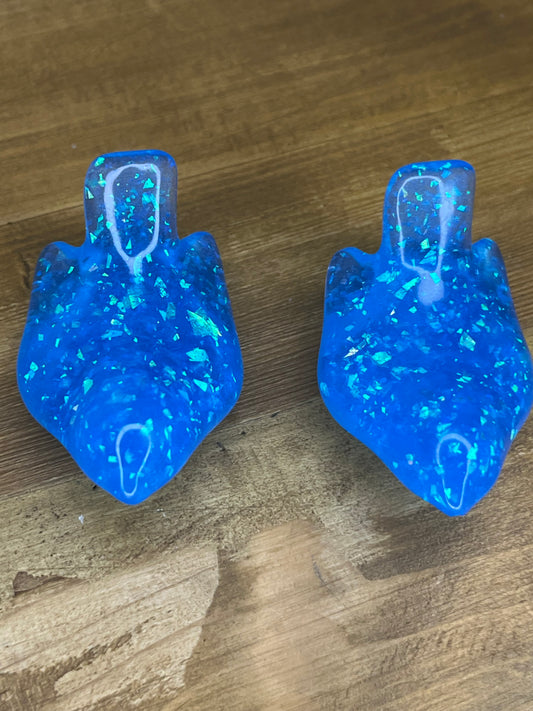 Two blue resin figurines with a teardrop shape and a textured, glittery appearance. They have a vibrant blue colour with embedded iridescent flakes, creating a sparkling effect. The background is a wooden surface.