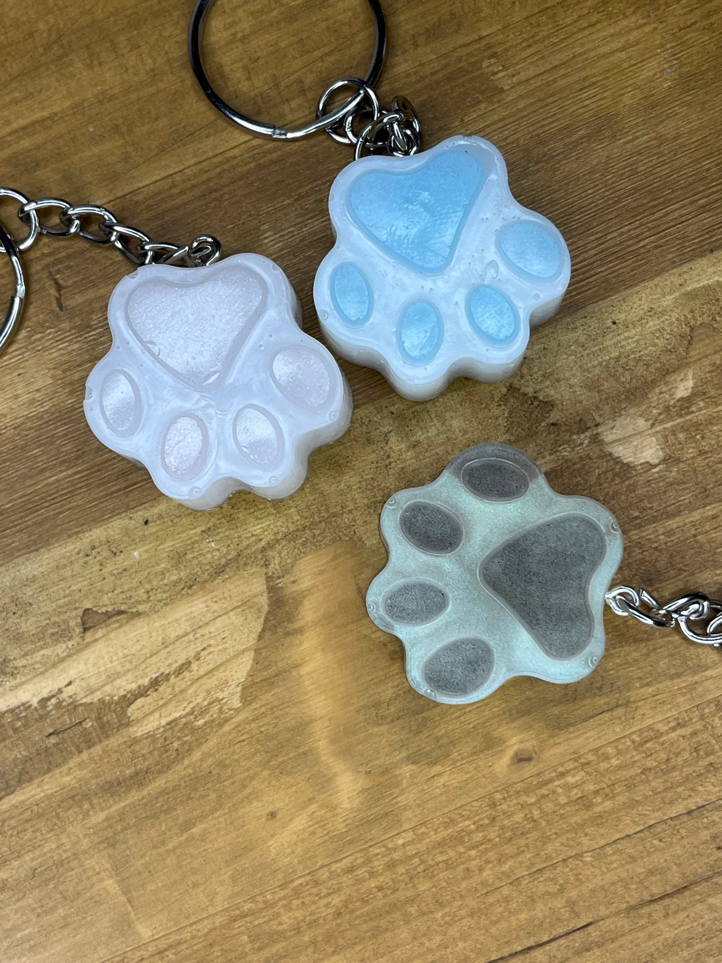 Three paw-shaped keyrings in light blue, pink, and grey. Each keyring has a silver chain and ring. The paw prints are made of a translucent material, with a glossy finish. The keyrings are on a wooden surface.