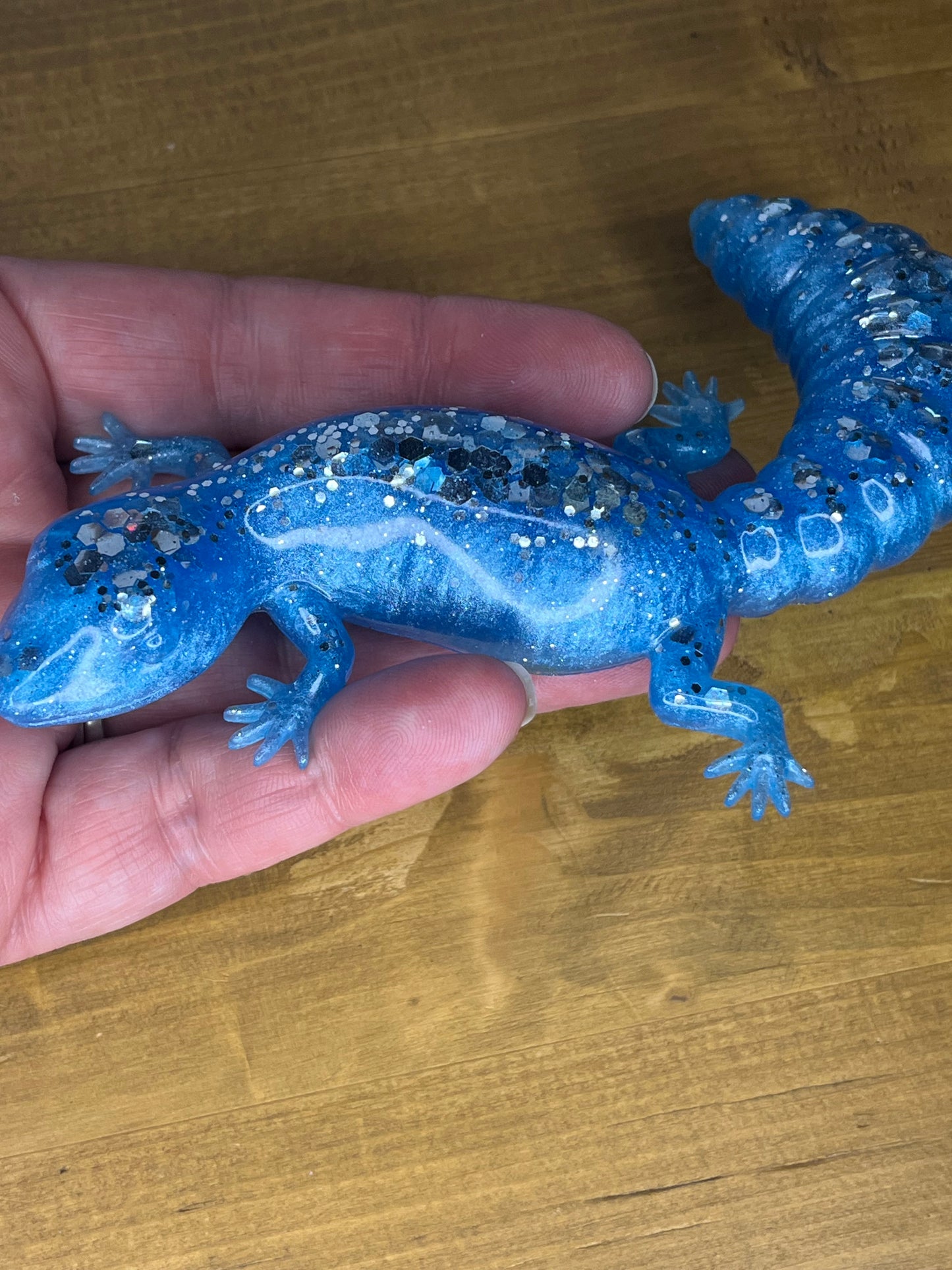 A blue resin lizard figurine with glitter accents. The lizard is posed with its legs outstretched and tail curved. The figurine is approximately 20 cm long.