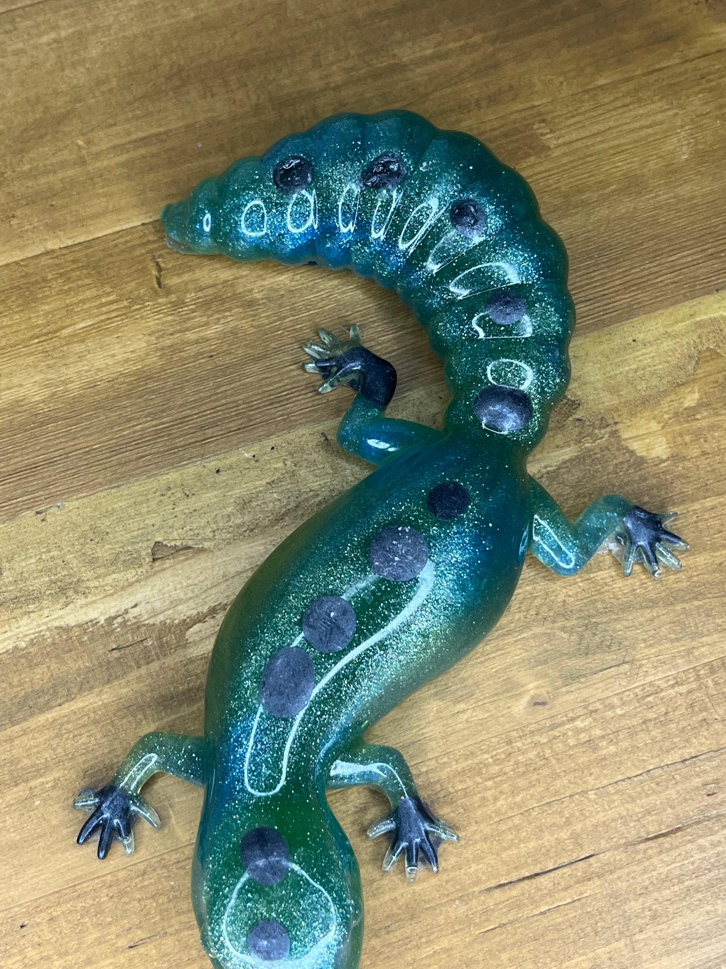 A teal and green salamander ornament with glitter and dark purple spots. The figurine has a curved tail and four tiny feet. It is displayed on a wooden surface.