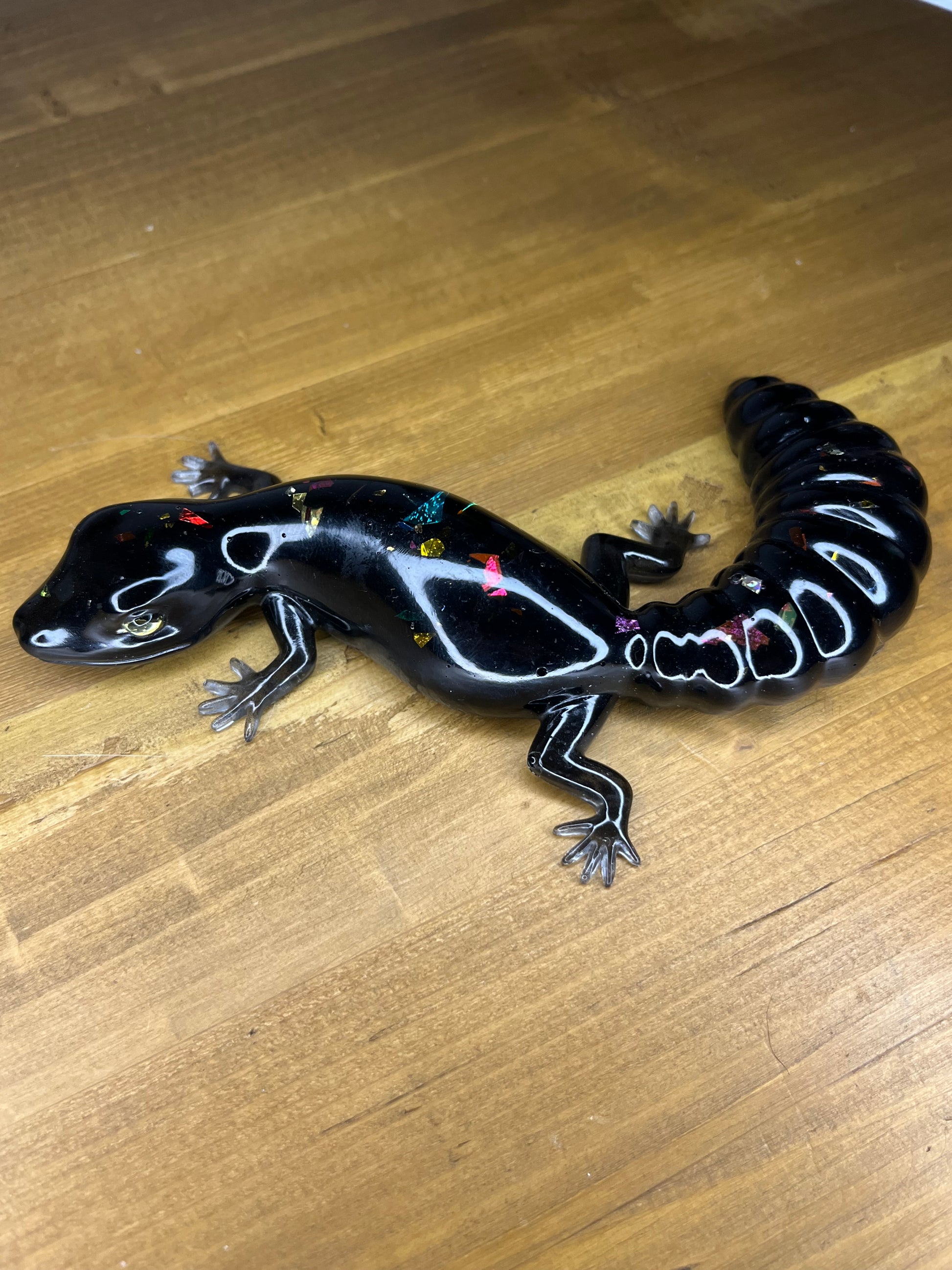 Glossy black resin salamander with colourful embedded glitter accents, displayed on a wooden surface.