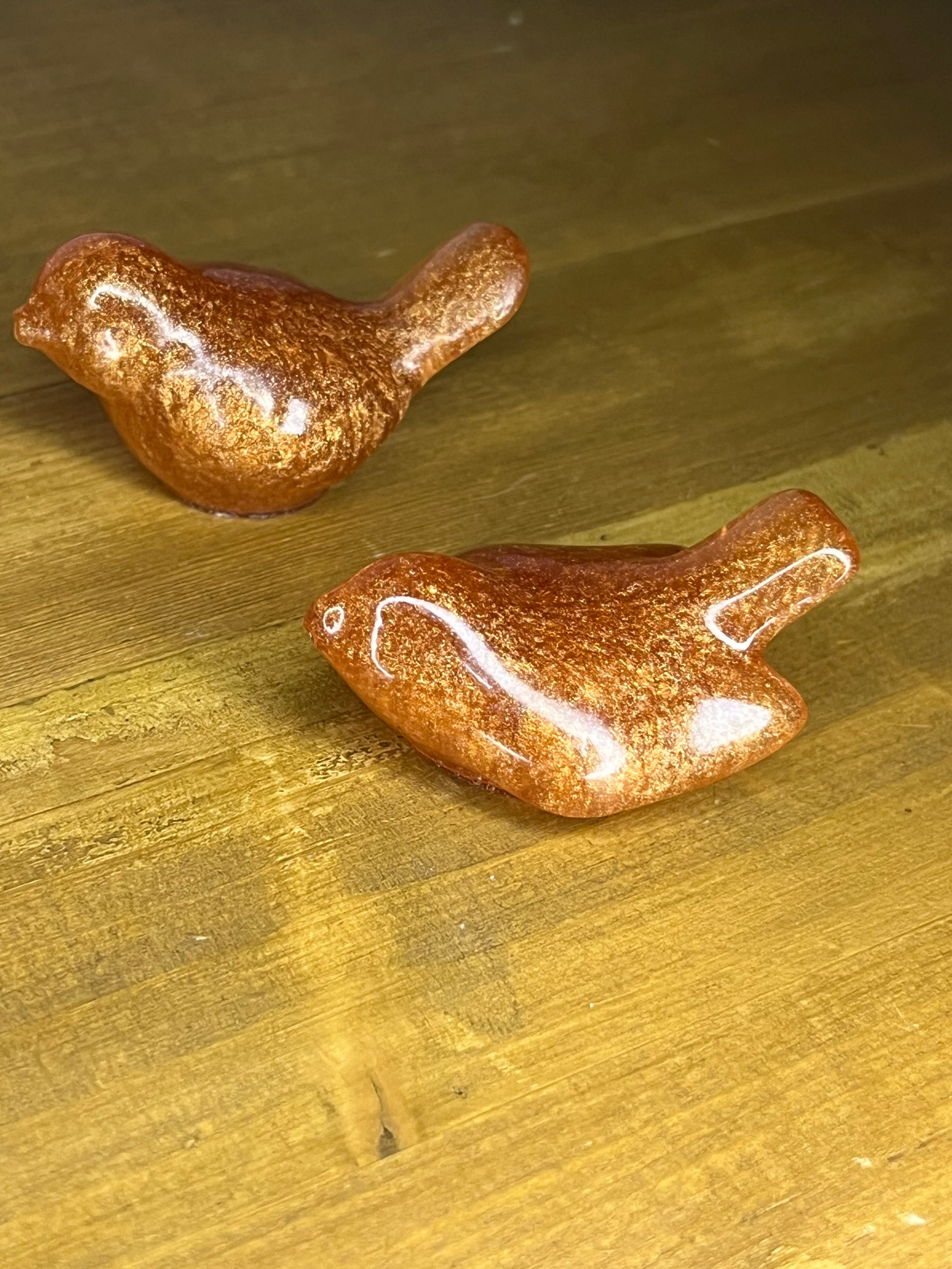 Two small, decorative bird figurines. The birds are a metallic copper colour with a glossy finish. They are positioned on a wooden surface with a warm, golden hue. The birds are detailed with wings and beaks.