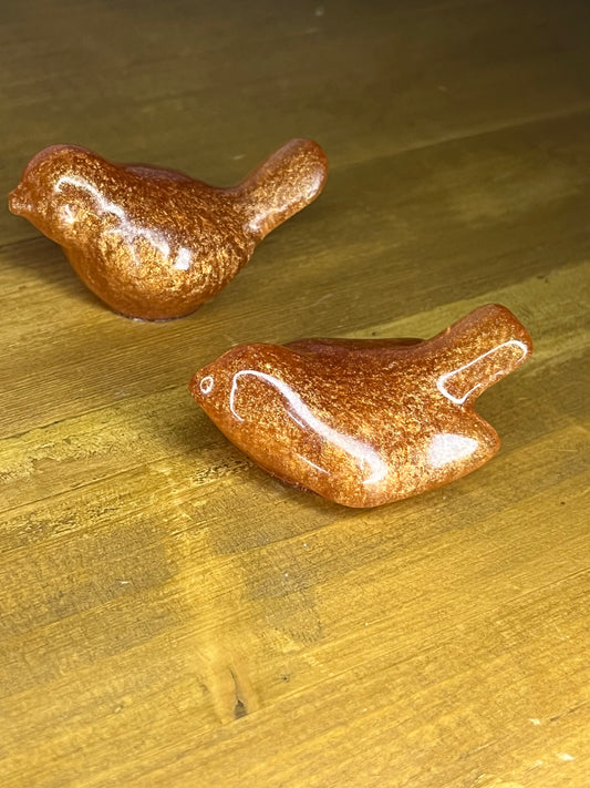 Two small, decorative bird figurines. The birds are a metallic copper colour with a glossy finish. They are positioned on a wooden surface with a warm, golden hue. The birds are detailed with wings and beaks.