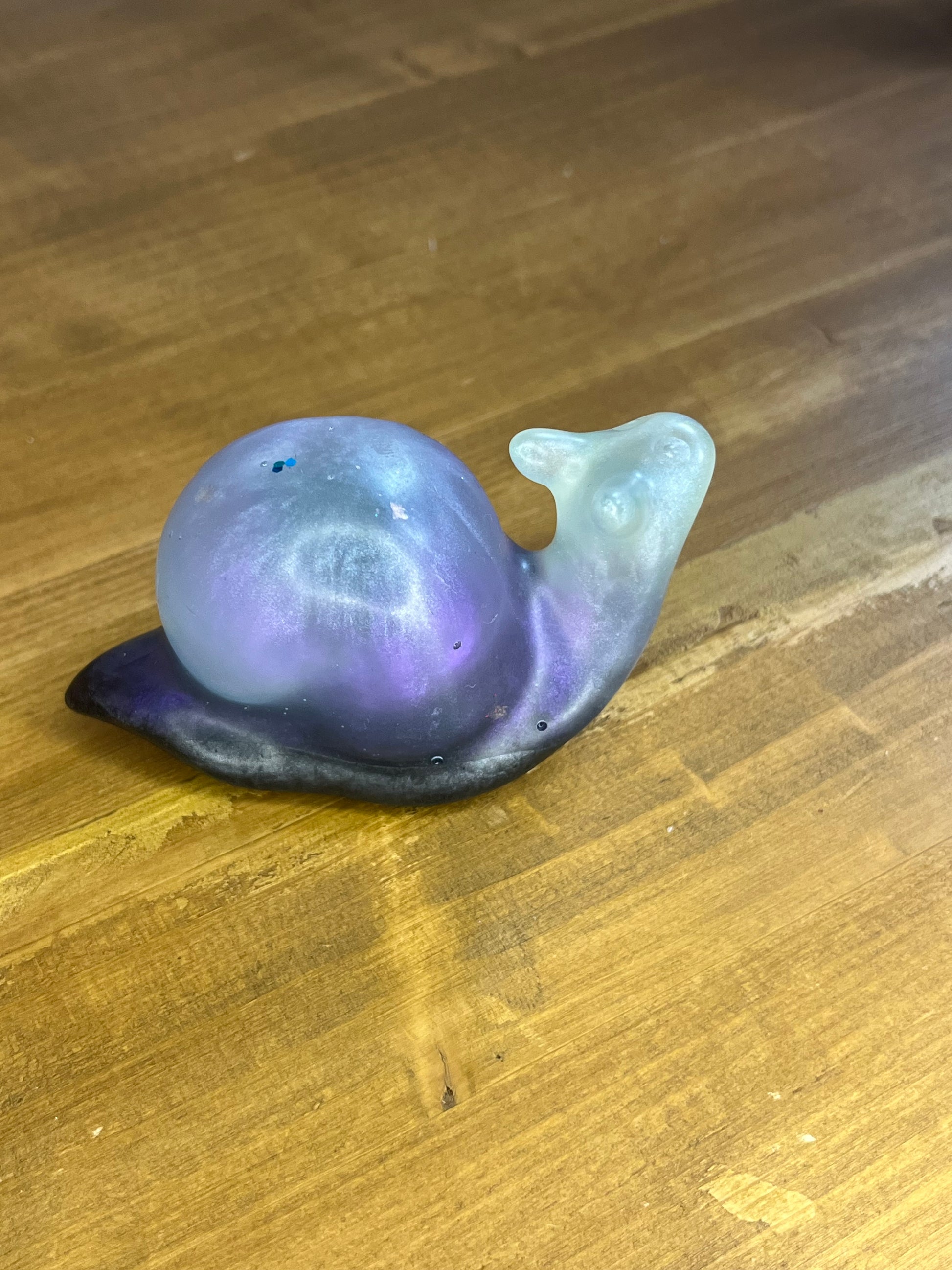 A decorative snail ornament with a colour gradient. The shell transitions from deep purple to a lighter, iridescent hue, with a black base. The snail's body is a translucent, light blue colour.