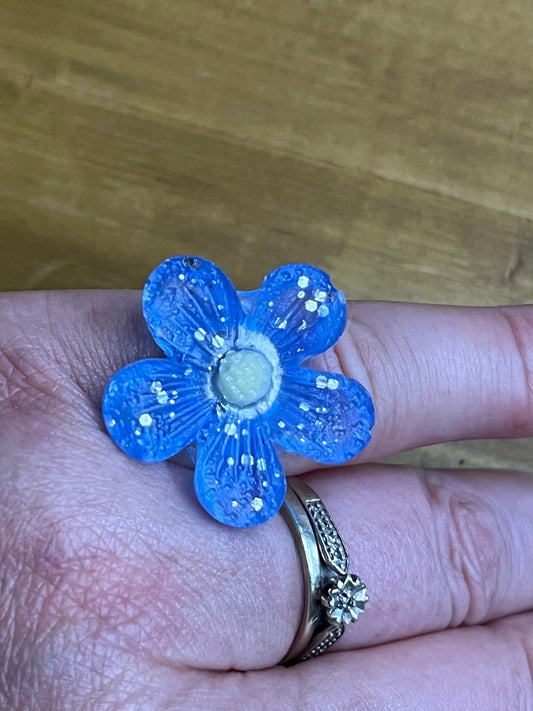 A blue flower-shaped ring with white glitter details and a pale yellow center, shown worn on a person's finger next to a silver ring.