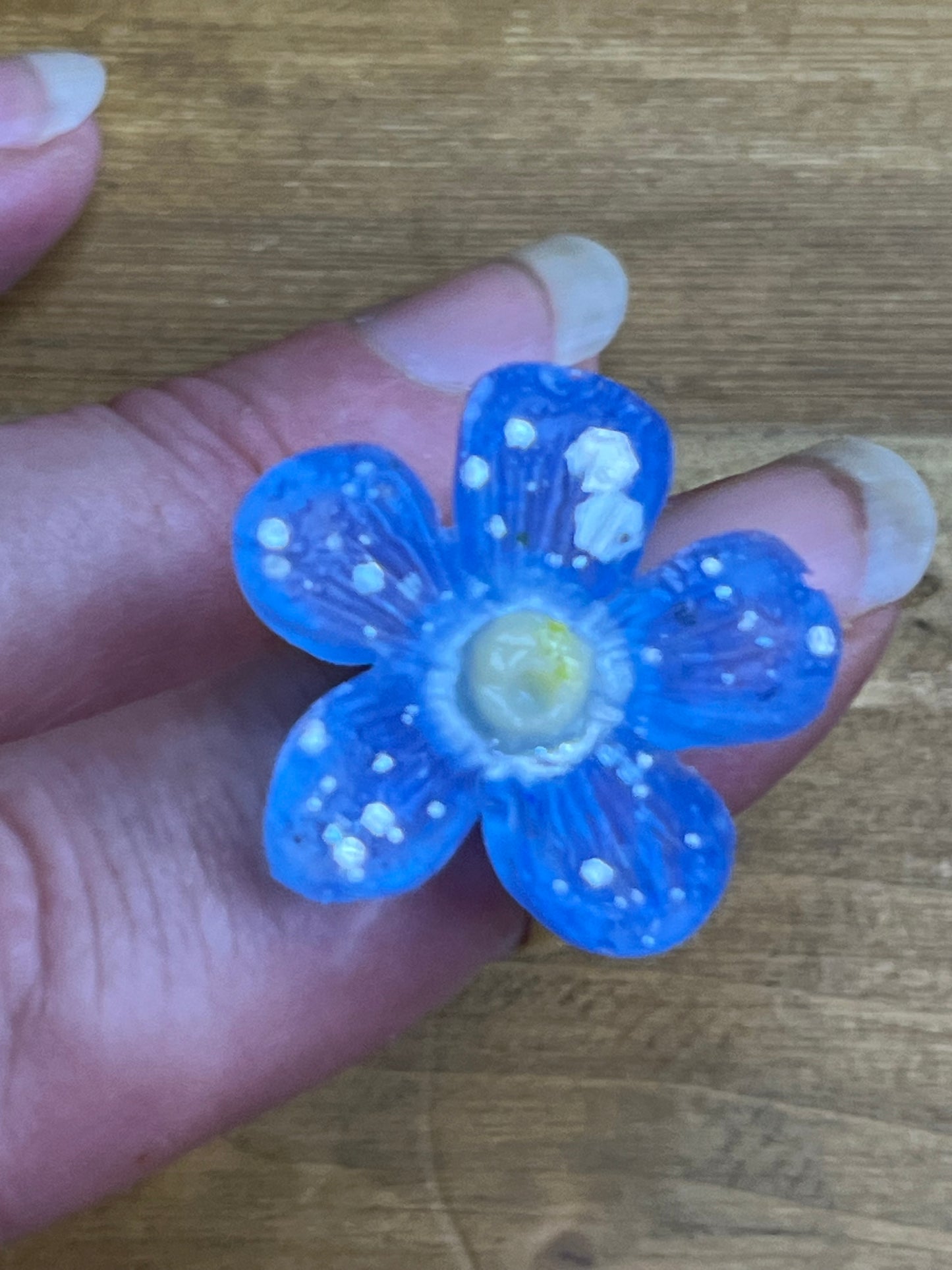 A handmade blue five-petal flower ring worn on a finger, decorated with white speckles and a yellow center, set against a wooden background.